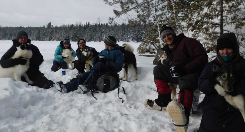 A group of people sit in the snow with sled dogs